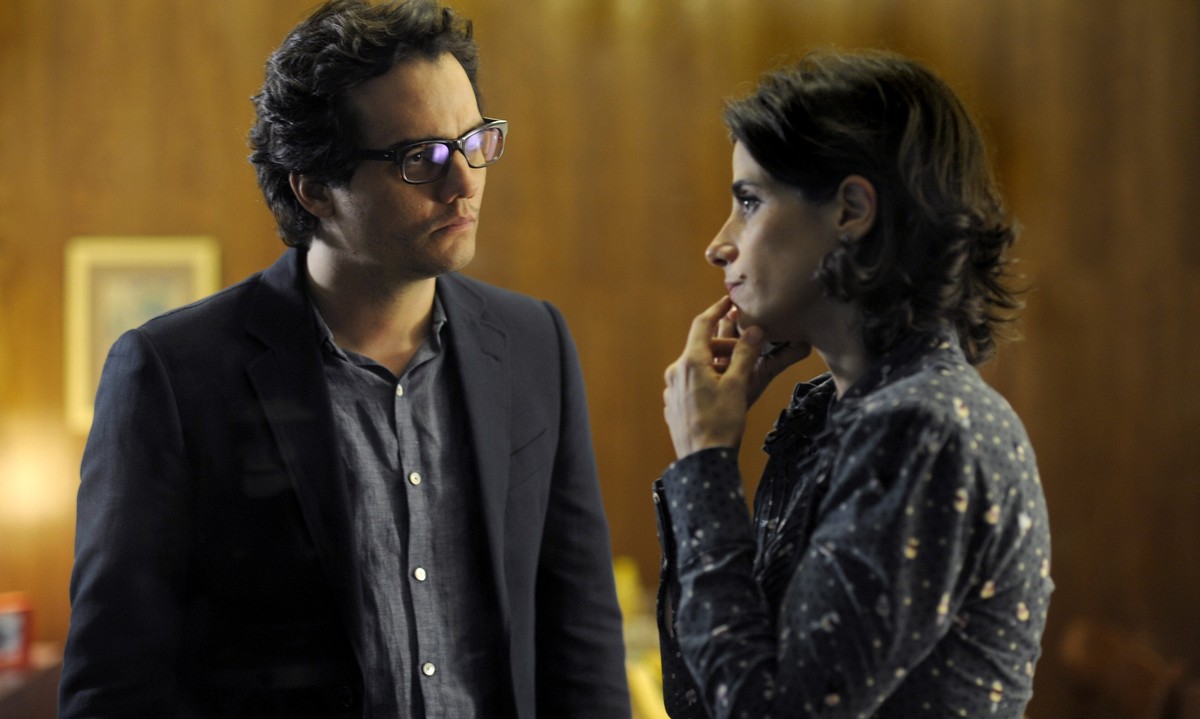 Wagner Moura, de óculos e blazer escuro, encara uma mulher pensativa em um escritório de paredes de madeira. Cena do filme A Busca (2012).