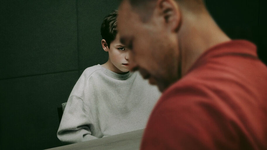 Onde assistir às séries vencedoras do Emmy 2025 8 Jamie Miller (Owen Cooper) encara com expressão séria ao fundo de uma sala de interrogatório, enquanto o pai aparece em primeiro plano desfocado, na série Adolescência, da Netflix.