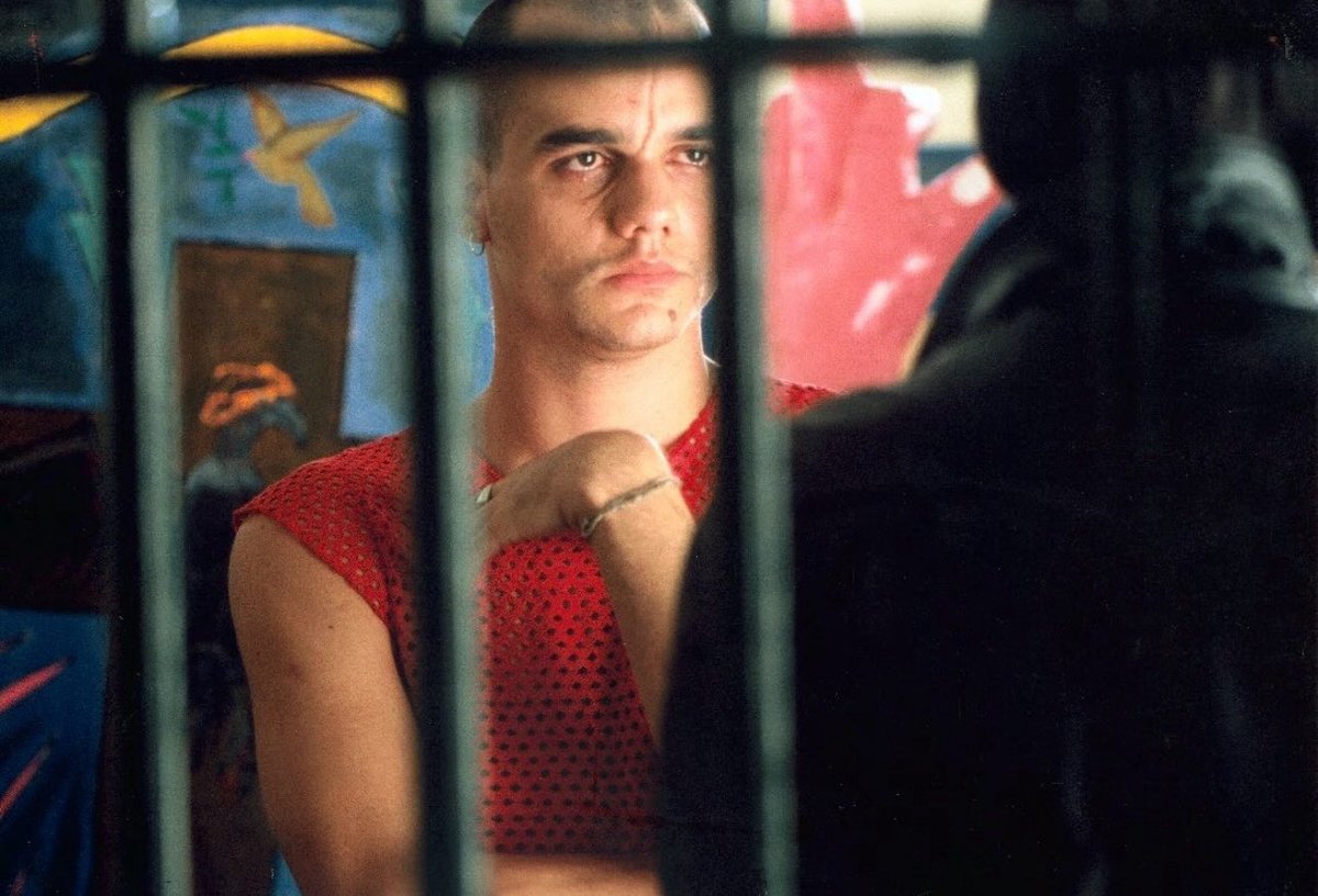 Wagner Moura com cabelo raspado e blusa vermelha rendada aparece atrás de grades, em um ambiente colorido, com expressão séria. Cena do filme As Três Marias (2002).