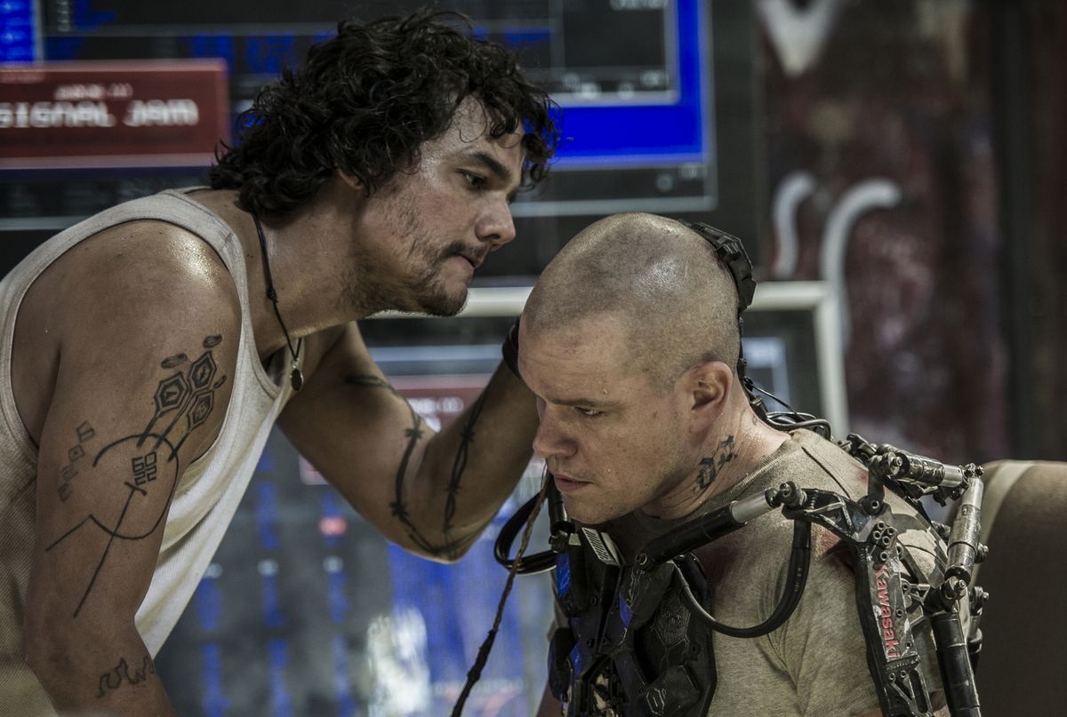 Wagner Moura, com cabelo desgrenhado, tatuagens e regata suja, segura Matt Damon pelo pescoço em um ambiente tecnológico. Cena do filme Elysium (2013).