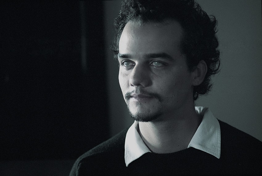 Wagner Moura com olhar perdido, pupilas esbranquiçadas e expressão sombria, veste camisa preta com gola branca. Cena do filme Nina (2004).