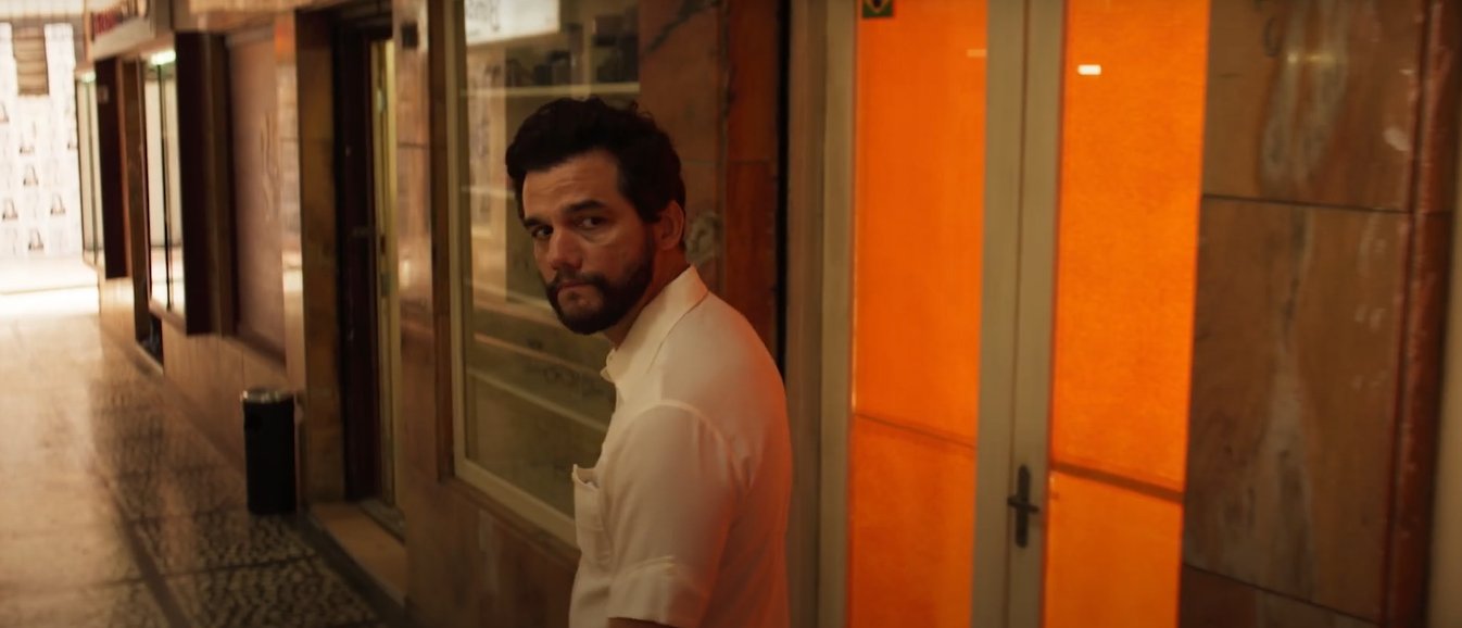 O ator Wagner Moura, de barba e camisa branca, olha para trás em um corredor estreito e pouco iluminado, próximo a uma porta com vidro laranja. Cena do filme O Agente Secreto (2025).