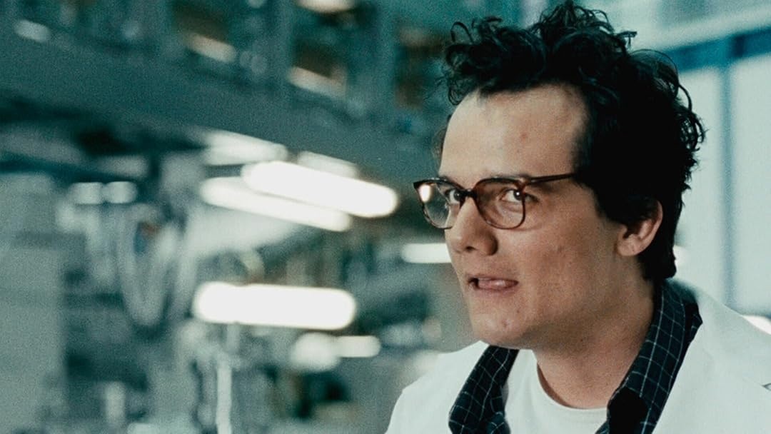 Wagner Moura, de óculos e jaleco branco sobre camisa xadrez, aparece em um laboratório moderno e iluminado, com expressão intensa. Cena do filme O Homem do Futuro (2011).
