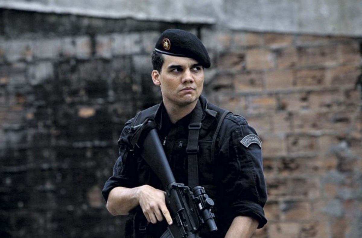 Wagner Moura caracterizado como Capitão Nascimento, em uniforme preto do BOPE e boina preta, segura um fuzil enquanto observa atentamente em uma favela. Cena do filme Tropa de Elite (2007).