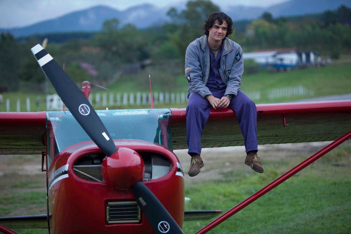 Wagner Moura, jovem e com cabelo encaracolado, veste macacão azul e casaco cinza, sentado sobre a asa de um pequeno avião vermelho em um campo aberto. Cena do filme Vips (2010).