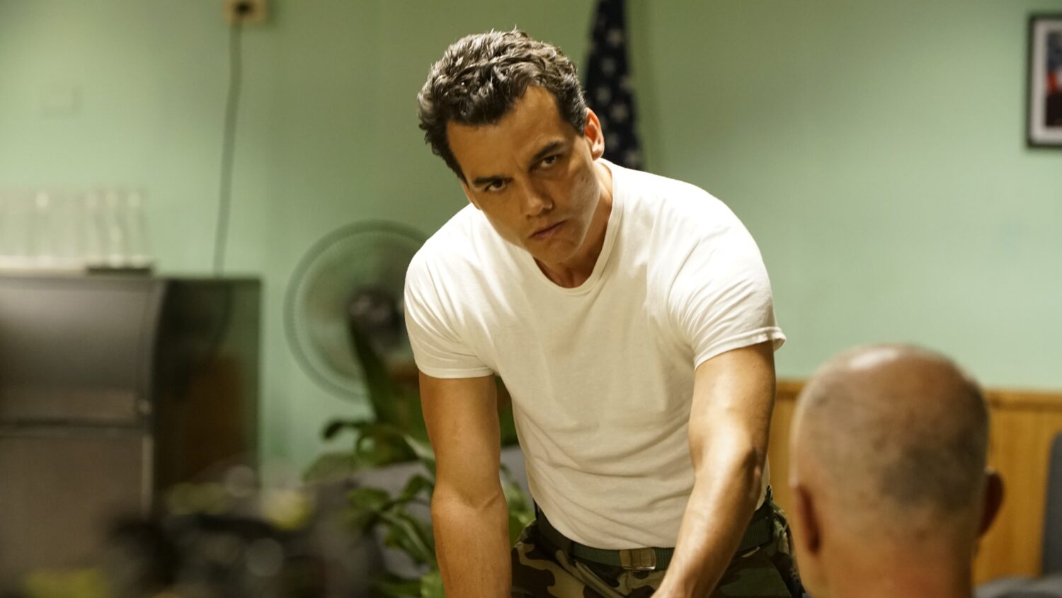 Wagner Moura, com cabelo curto e expressão séria, usa camiseta branca ajustada e calça camuflada enquanto encara outro personagem em uma sala. Cena do filme Wasp Network (2019).