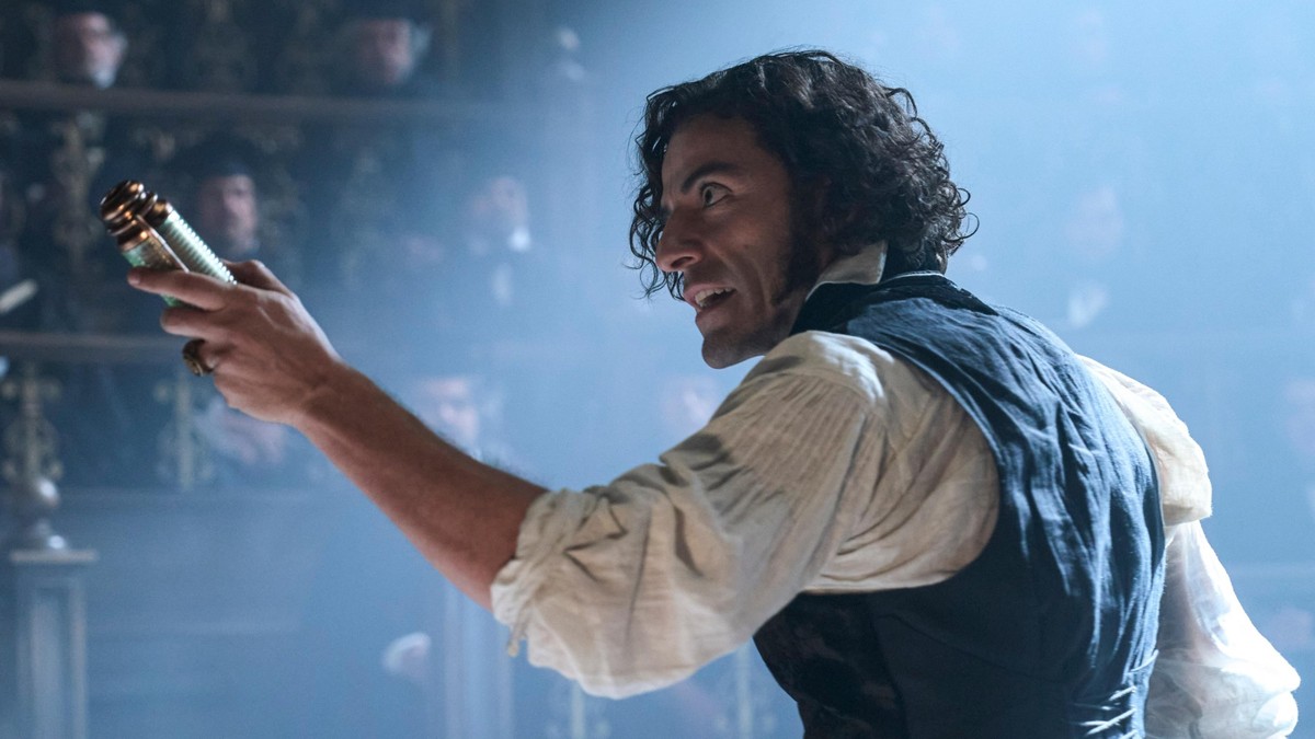 Cena do filme Frankenstein (2026), dirigido por Guillermo del Toro. O ator Oscar Isaac, no papel do Dr. Victor Frankenstein, aparece em um ambiente acadêmico iluminado por luz difusa, vestindo camisa branca e colete escuro. Ele segura um objeto cilíndrico metálico, com expressão intensa e olhar fixo, enquanto discursa diante de uma plateia ao fundo.