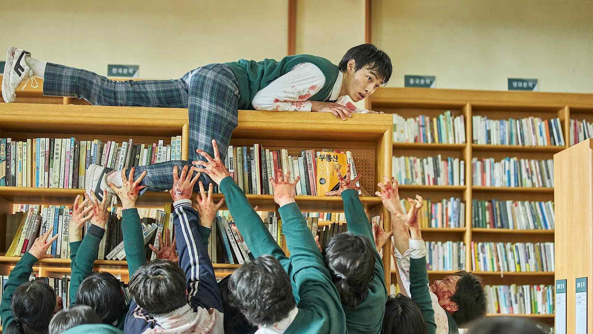 Cena de All of Us Are Dead (2022–) mostra um estudante em cima de uma estante de livros tentando escapar de um grupo de zumbis que o alcança com as mãos ensanguentadas.
