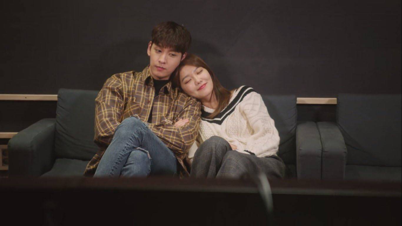 Cena de So I Married the Anti-Fan (2021) mostra um casal sentado em um sofá; a mulher sorri encostando a cabeça no ombro do homem, em um momento de ternura.