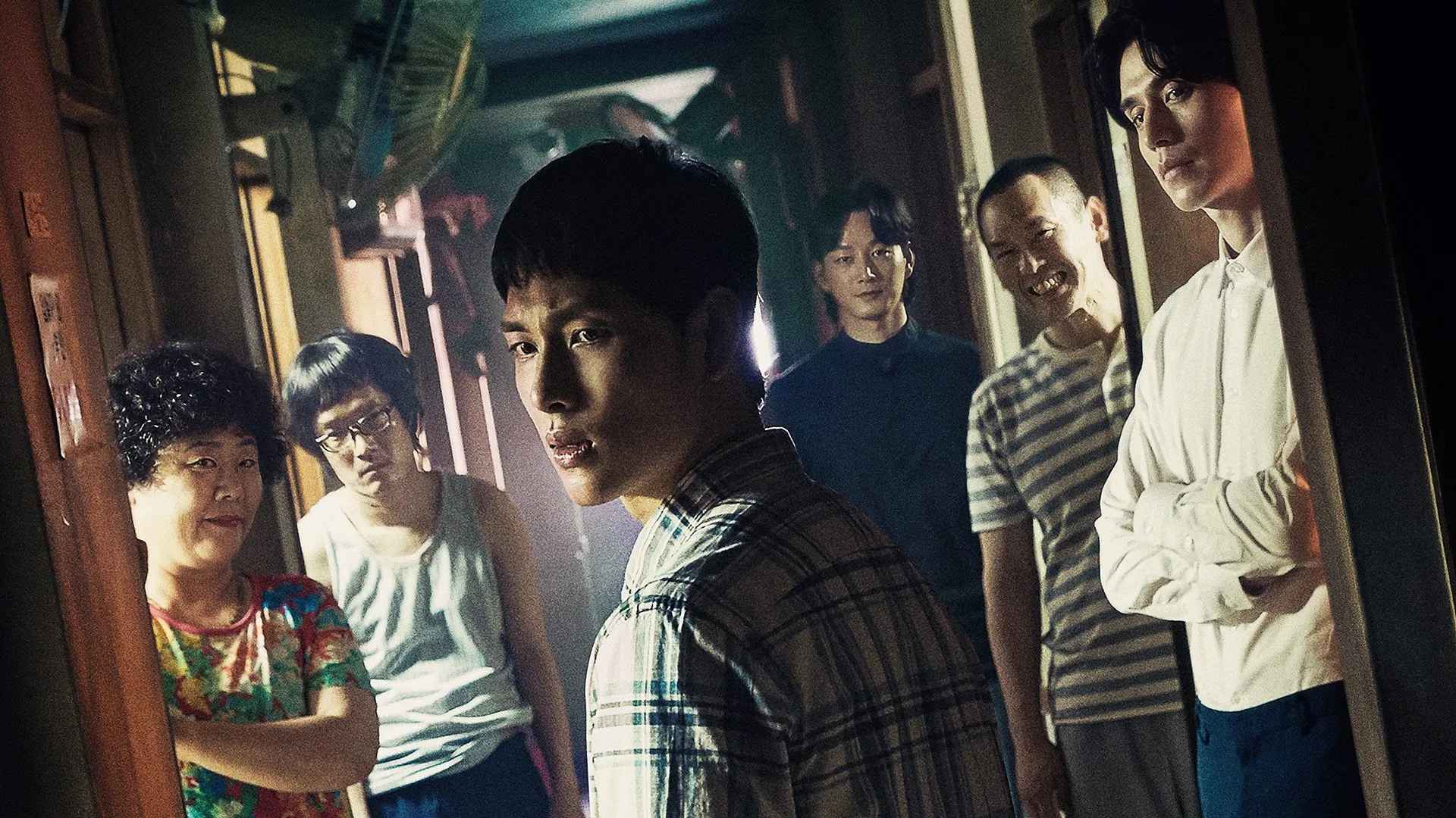 Cena de Strangers from Hell (2019) mostra um jovem de expressão tensa cercado por vizinhos com olhares sombrios em um corredor mal-iluminado de um prédio.