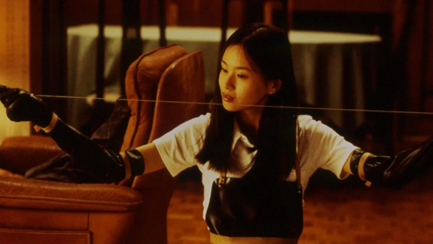 Filmes de terror asiáticos famosos: 12 obras que conquistaram o mundo 1 mulher com luvas pretas segura um fio metálico esticado, prestes a agir, em uma sala iluminada por luz amarela.