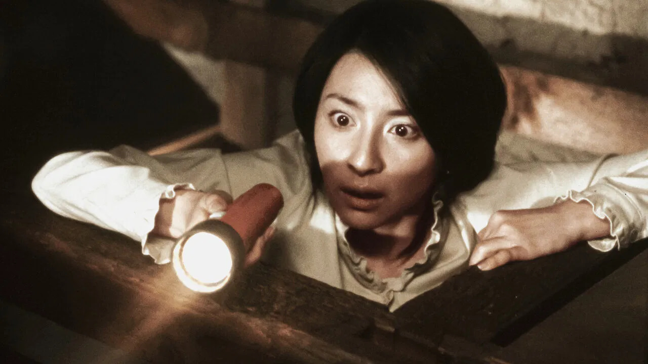 Filmes de terror asiáticos famosos: 12 obras que conquistaram o mundo 4 mulher segura uma lanterna e observa algo com expressão de espanto em um ambiente escuro de madeira.