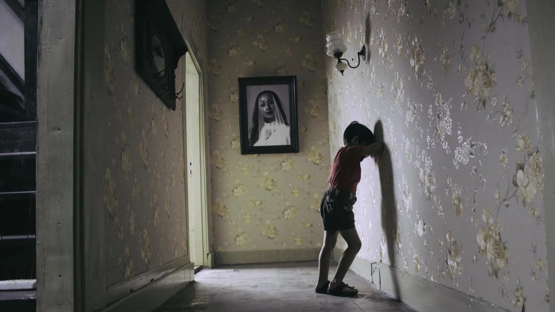 Filmes de terror asiáticos famosos: 12 obras que conquistaram o mundo 11 menino encosta a cabeça na parede de um corredor antigo decorado com papel floral e um retrato emoldurado ao fundo.