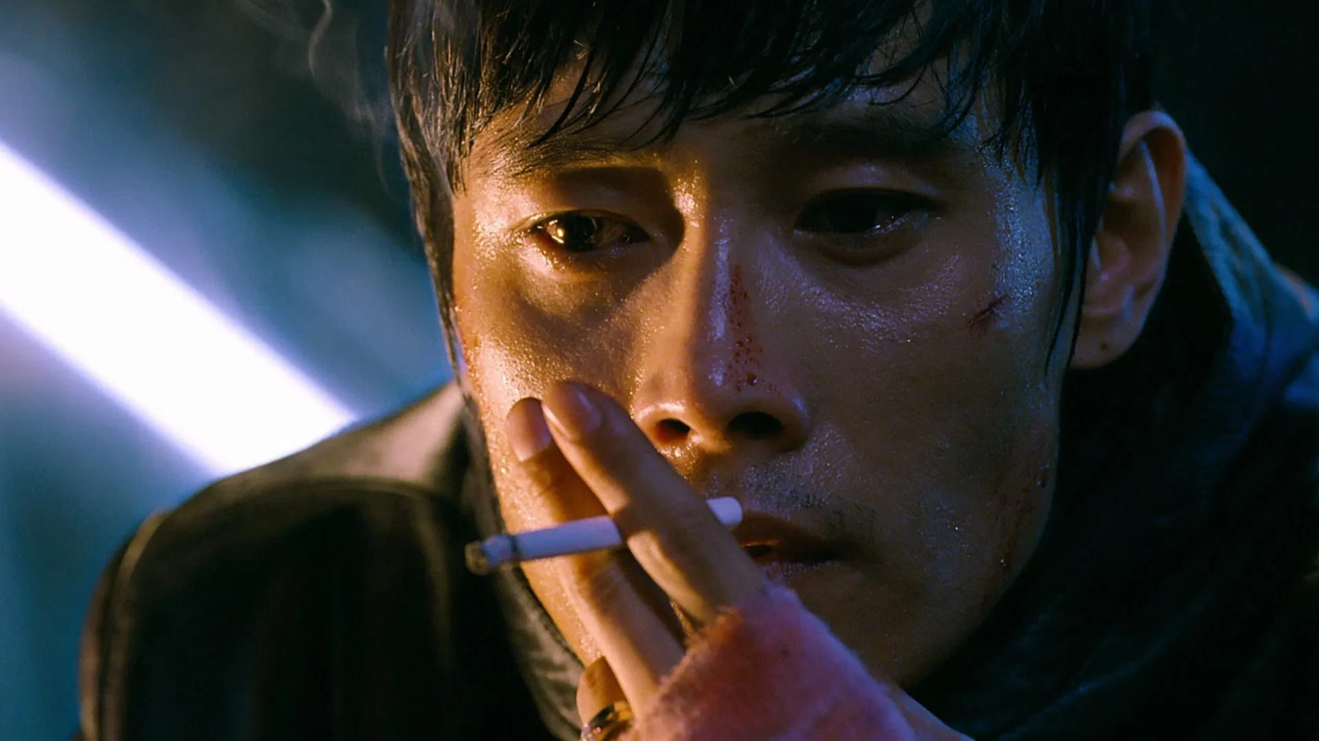 Filmes de terror asiáticos famosos: 12 obras que conquistaram o mundo 6 homem com expressão abatida e ferimentos no rosto segura um cigarro aceso, iluminado por uma luz fria que acentua o clima de tensão.