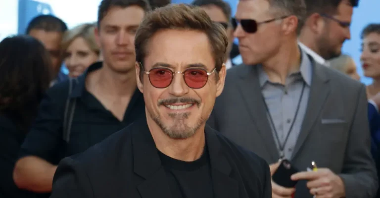 Robert Downey Jr. sorri usando óculos vermelhos durante evento da Marvel em tapete vermelho.